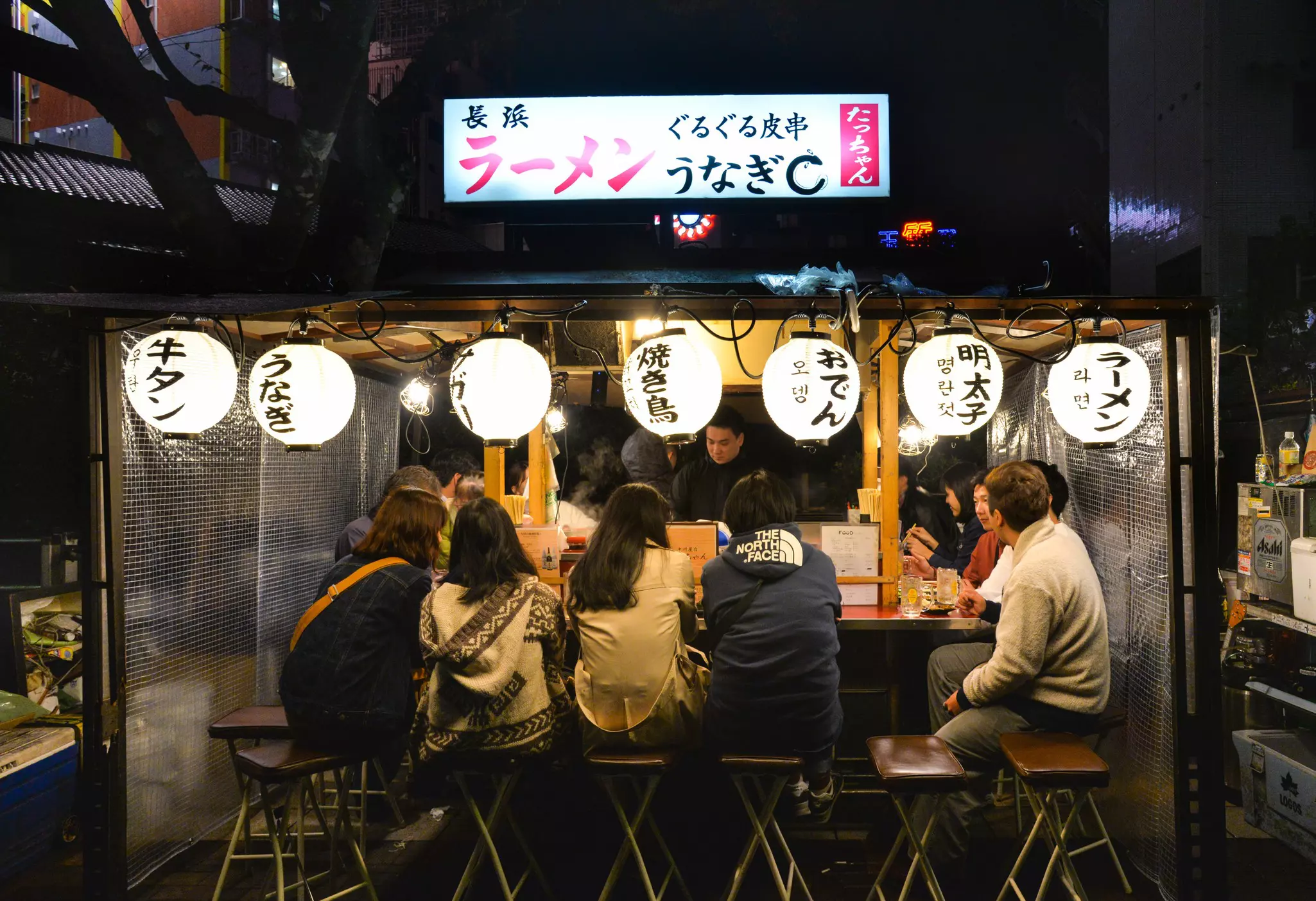 Diners at a counter at a street food stall. White lanterns with Japanese writing hang above them.