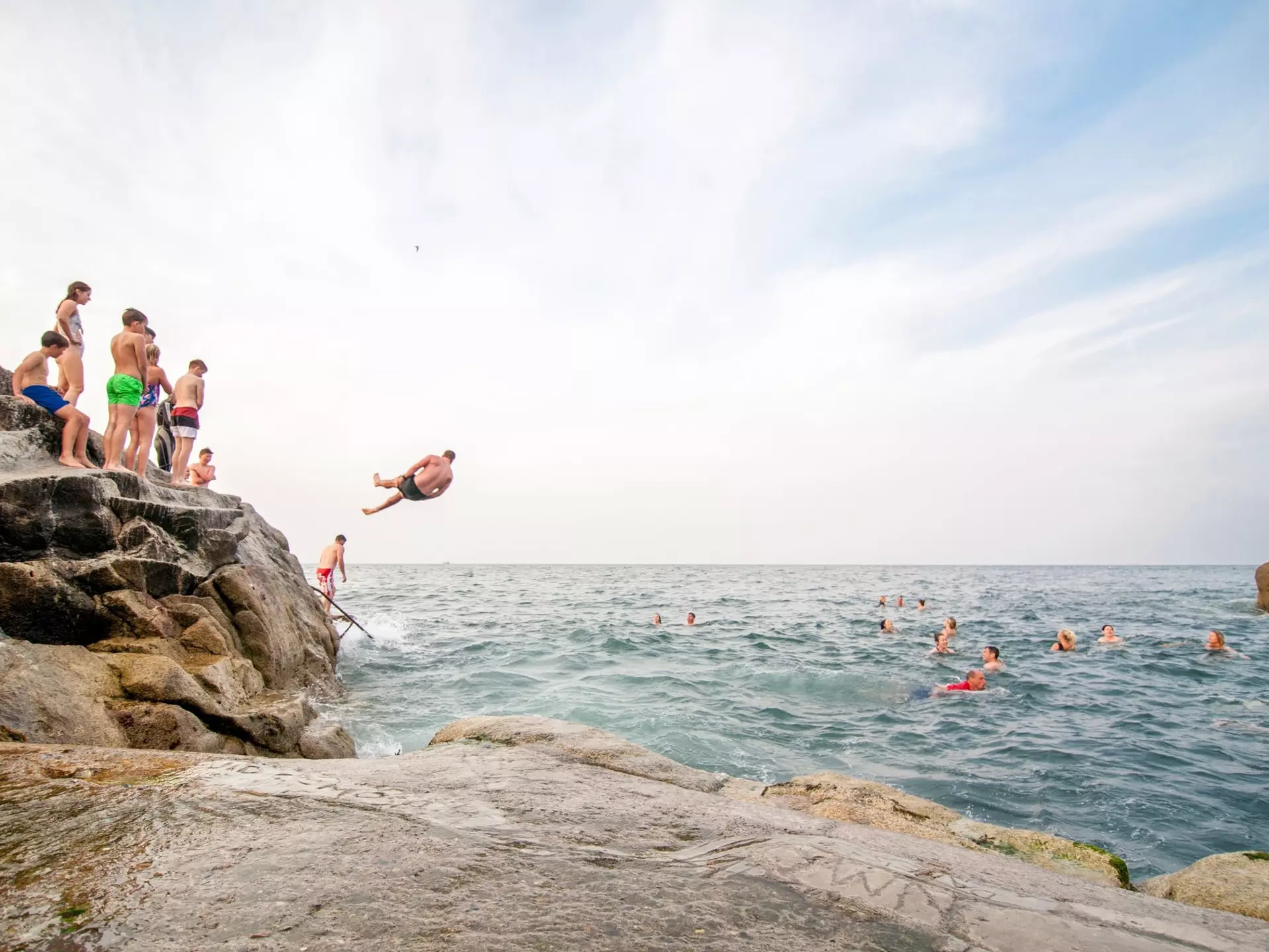 People line up to jump into the water from a small cliff. Other bathers are pictured swimming in a cove off the sea.