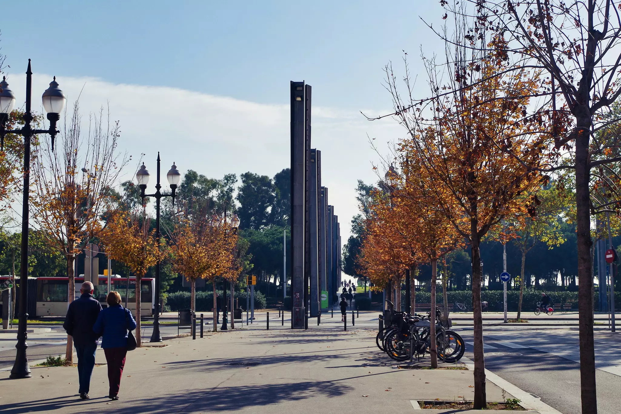 People walking down Rambla del Poblenou; the esplanade is lined with ornate lamp-posts and trees in autumnal colours, and there are sculptural columns further along the Rambla.