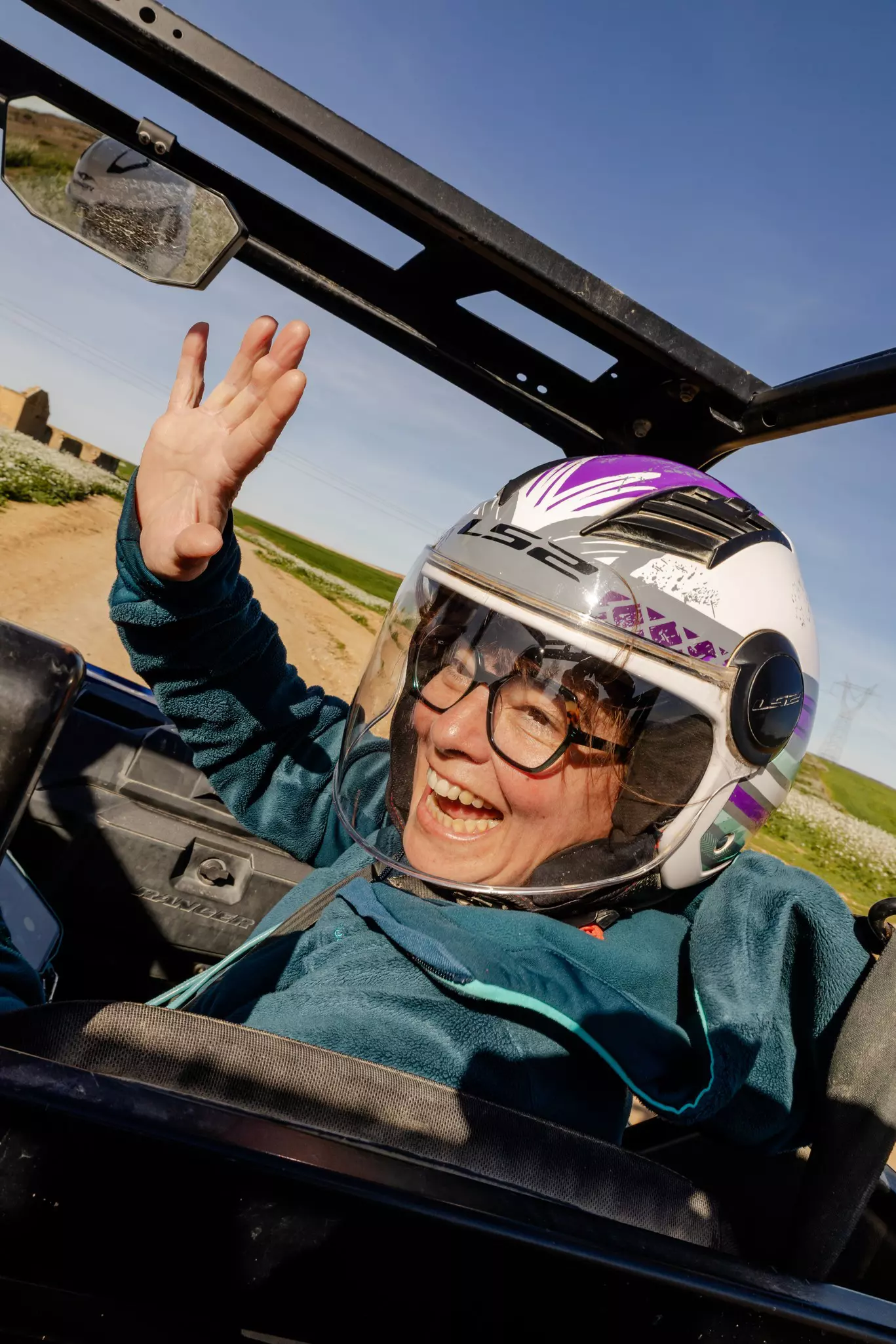 A woman wearing a safety helmet smiles and waves as she travels along in the front passenger seat of an open-top buggy.