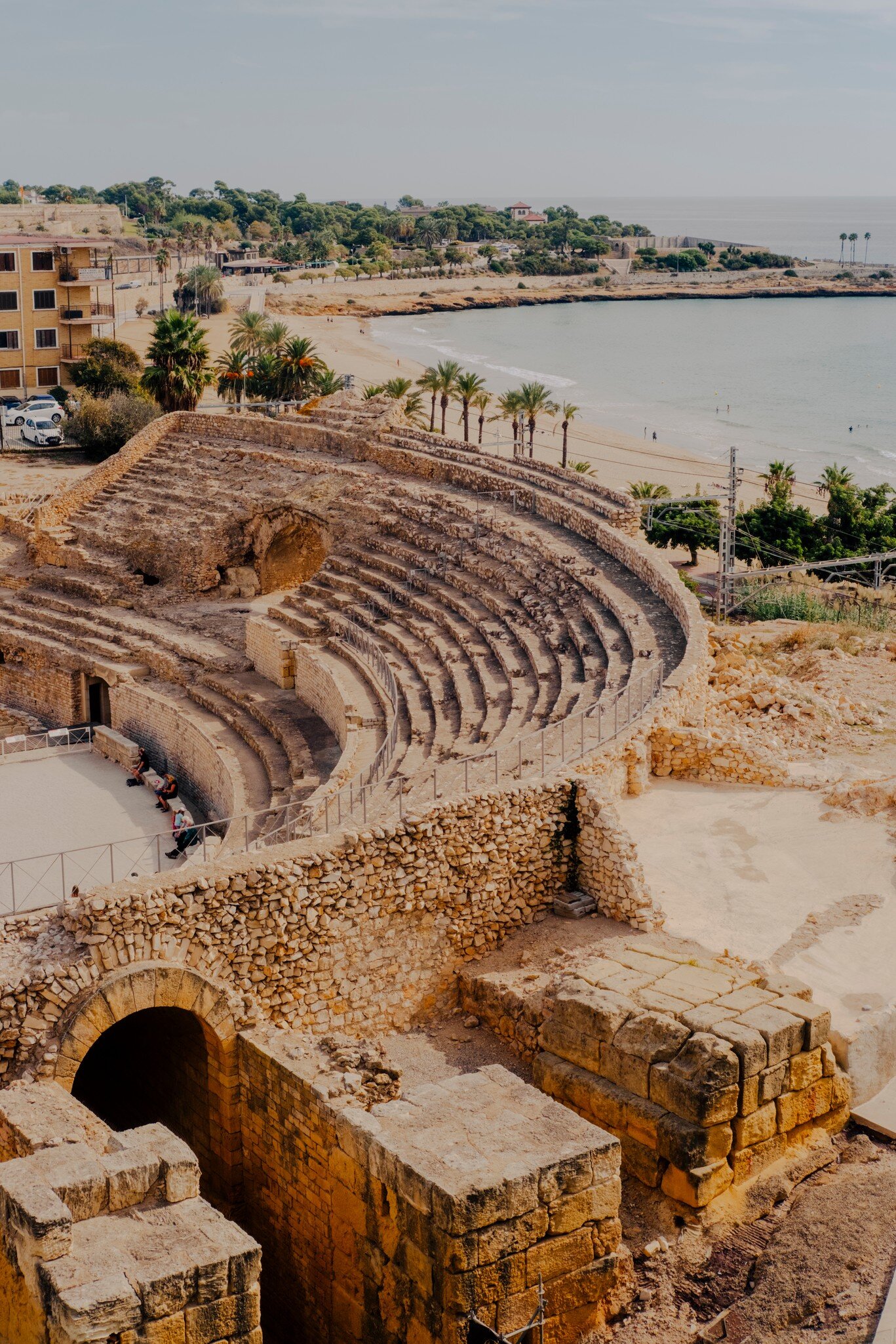 Tiered stone seating of an ancient amphitheatre above a sandy beach lined with palm trees and calm sea.