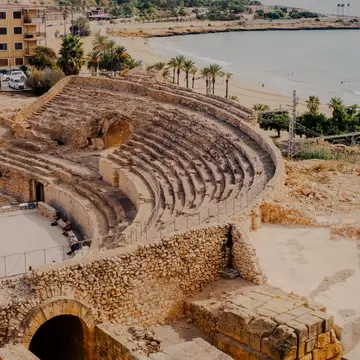 Tiered stone seating of an ancient amphitheatre above a sandy beach lined with palm trees and calm sea.
