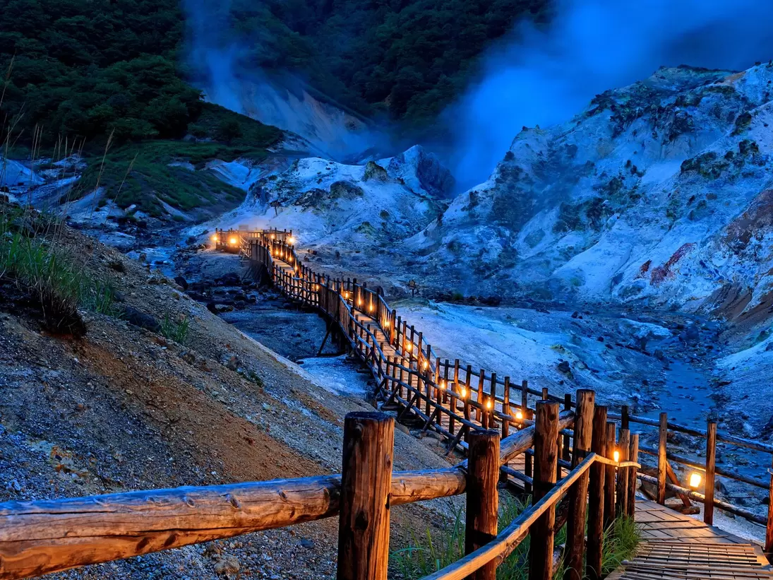 An illuminated wooden boardwalk through a volcanic landscape at dusk.