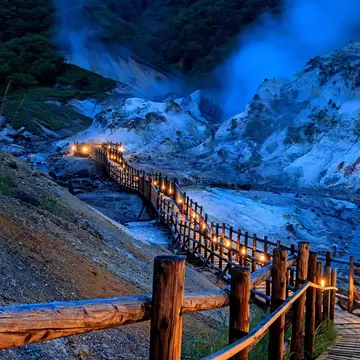 An illuminated wooden boardwalk through a volcanic landscape at dusk.
