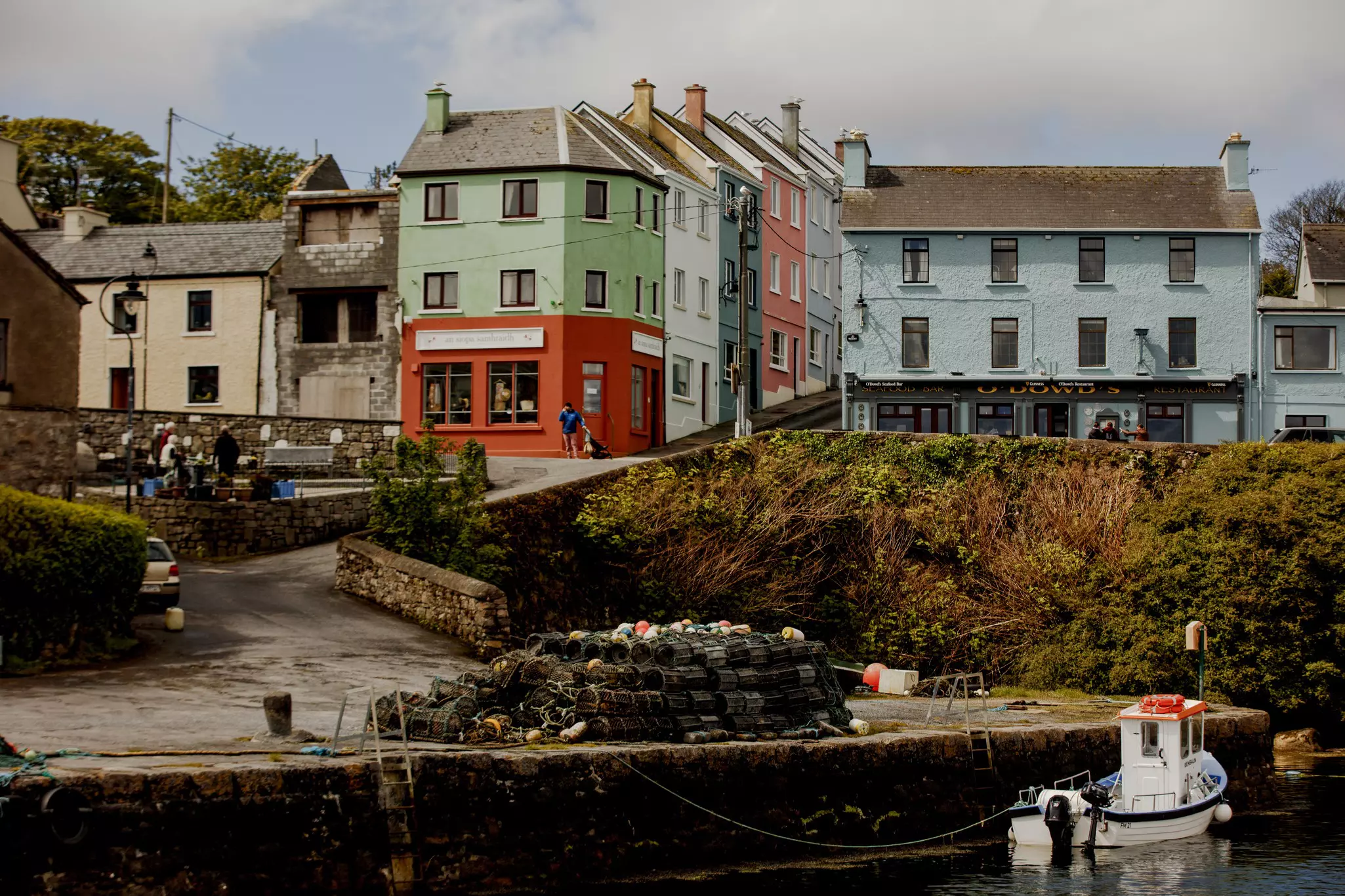 A harborfront village with pubs and shops facing the sea. Groups of people sit on benches by the water.