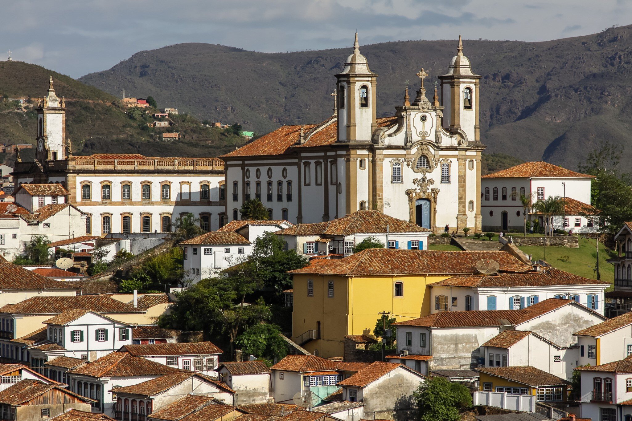 A white twin-towered baroque church stands above terracotta-roofed colonial houses, with green hills behind.