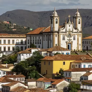 A white twin-towered baroque church stands above terracotta-roofed colonial houses, with green hills behind.