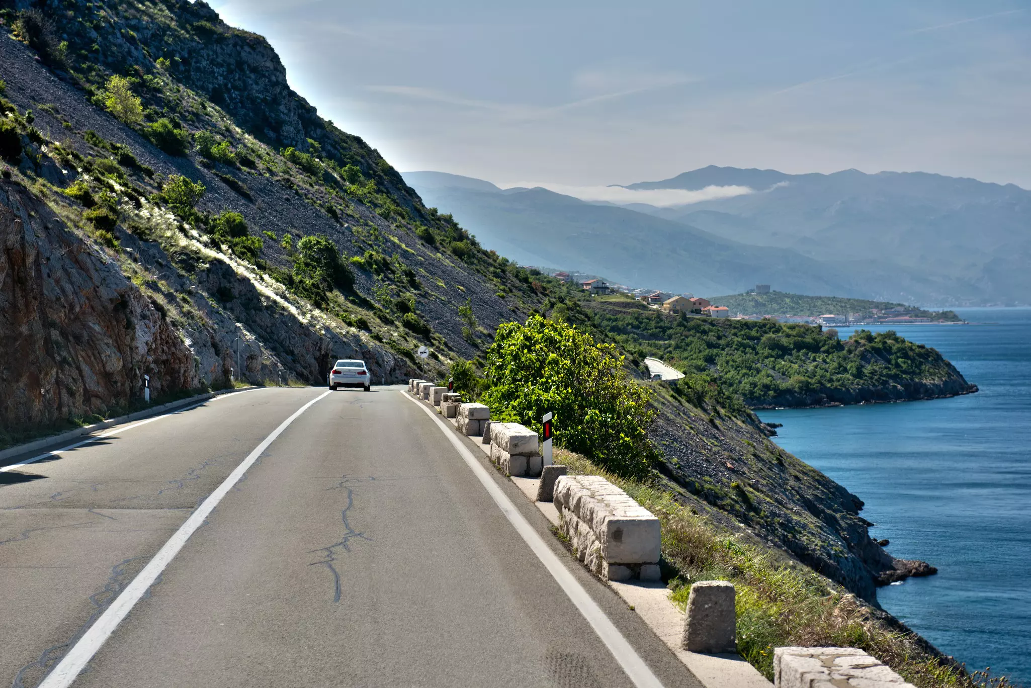 A car is seen from behind on a road hugging a coast, with a steep hill rising to the left.