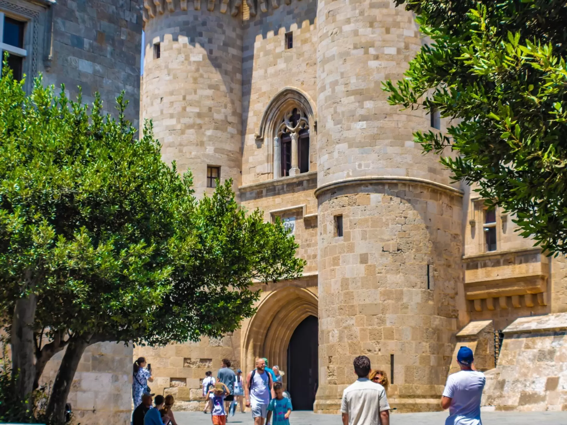 Entrance to a large sandstone castle with people gathered around a tree outside the walls