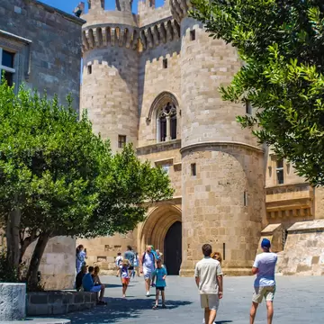 Entrance to a large sandstone castle with people gathered around a tree outside the walls