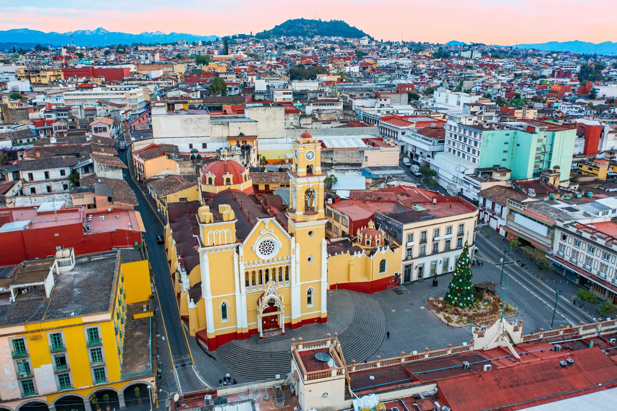 Aerial view of a historic city with a bold yellow building in the foreground and red rooftops spreading into the distance. A mountain and hillsides are on the far horizon.