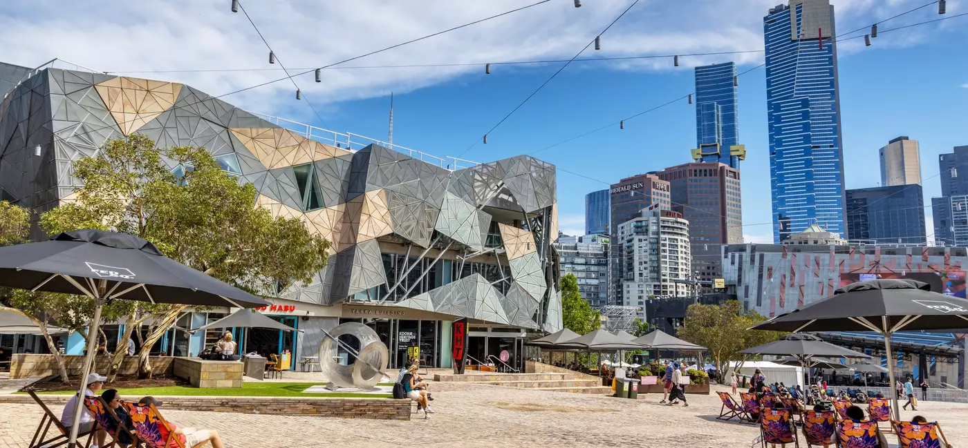 People sitting on lounge chairs under umbrellas on a brick patio by a low angular building.