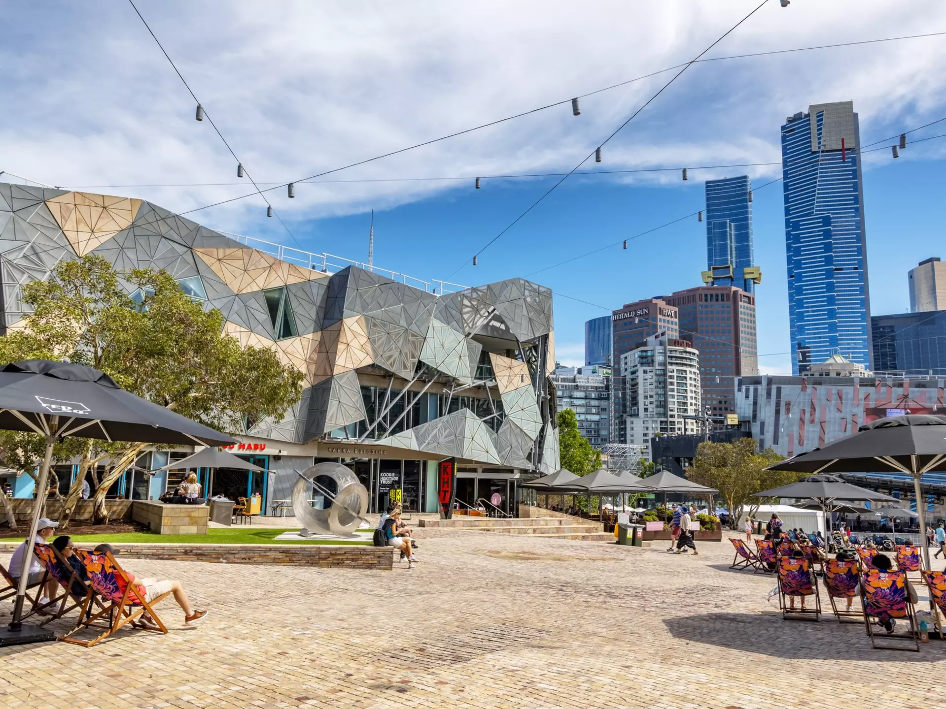 People sitting on lounge chairs under umbrellas on a brick patio by a low angular building.