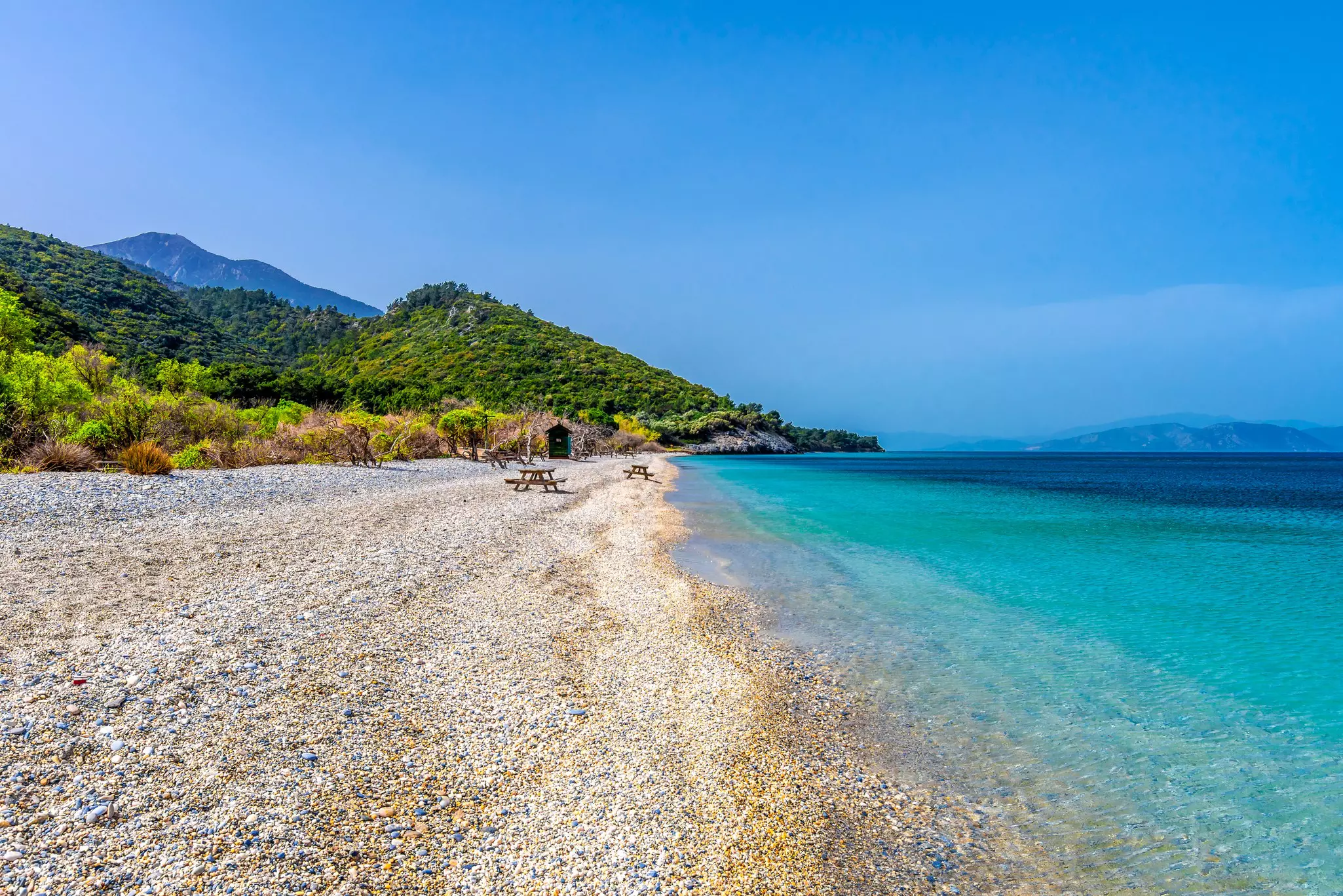 An empty pebbly beach in Turkey lapped by turquoise ocean. Several wooden picnic benches dot the shoreline.