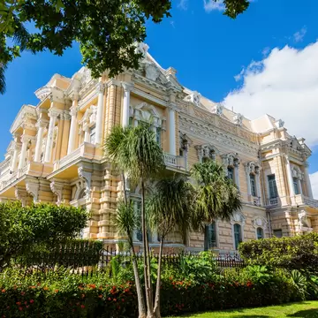 Cream-colored mansion with columns, carved balustrades and palms in the garden in front, under a blue sky.
