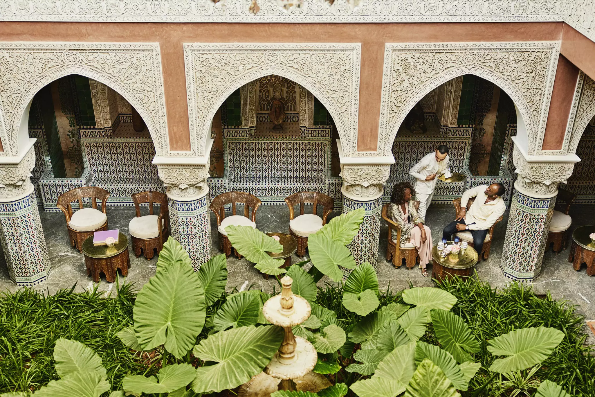 Extreme wide high angle shot of couple being served tea by waiter in courtyard of luxury hotel while on vacation in Marrakesh, Morocco
