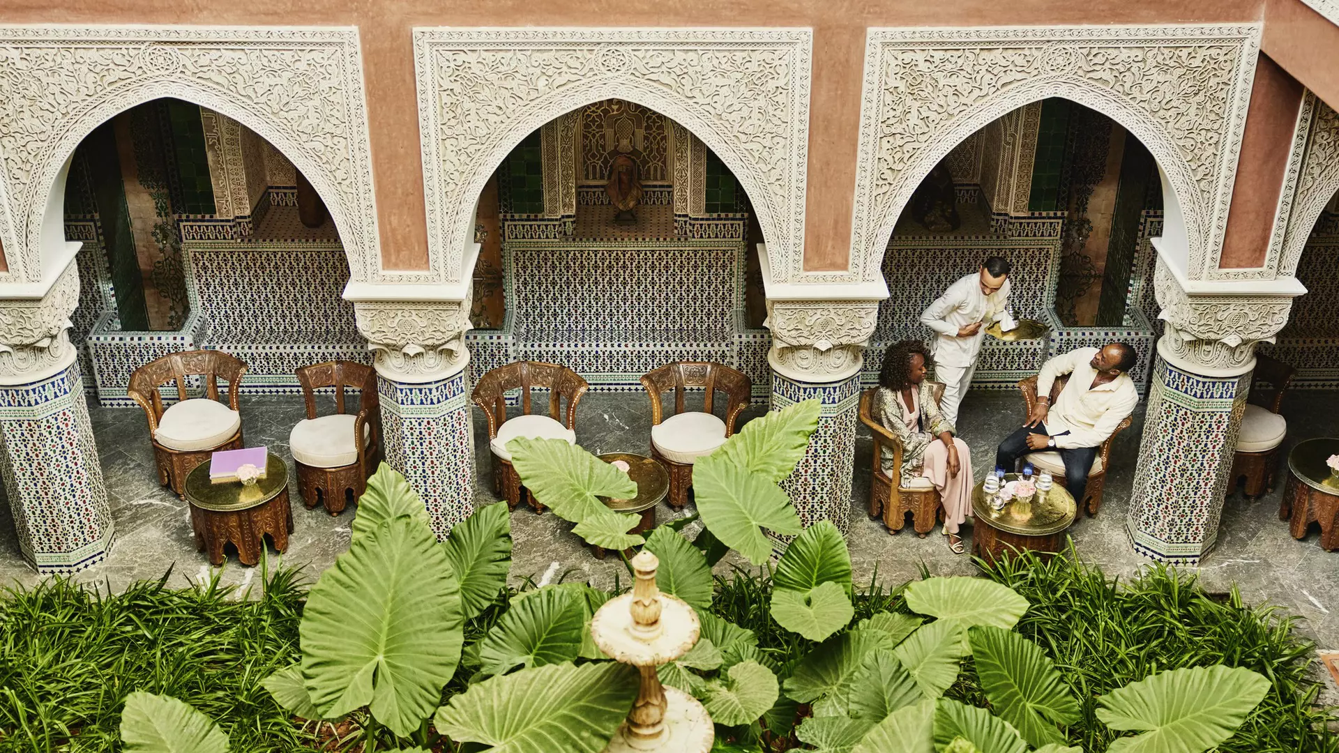Extreme wide high angle shot of couple being served tea by waiter in courtyard of luxury hotel while on vacation in Marrakesh, Morocco © Thomas Barwick / Getty Images