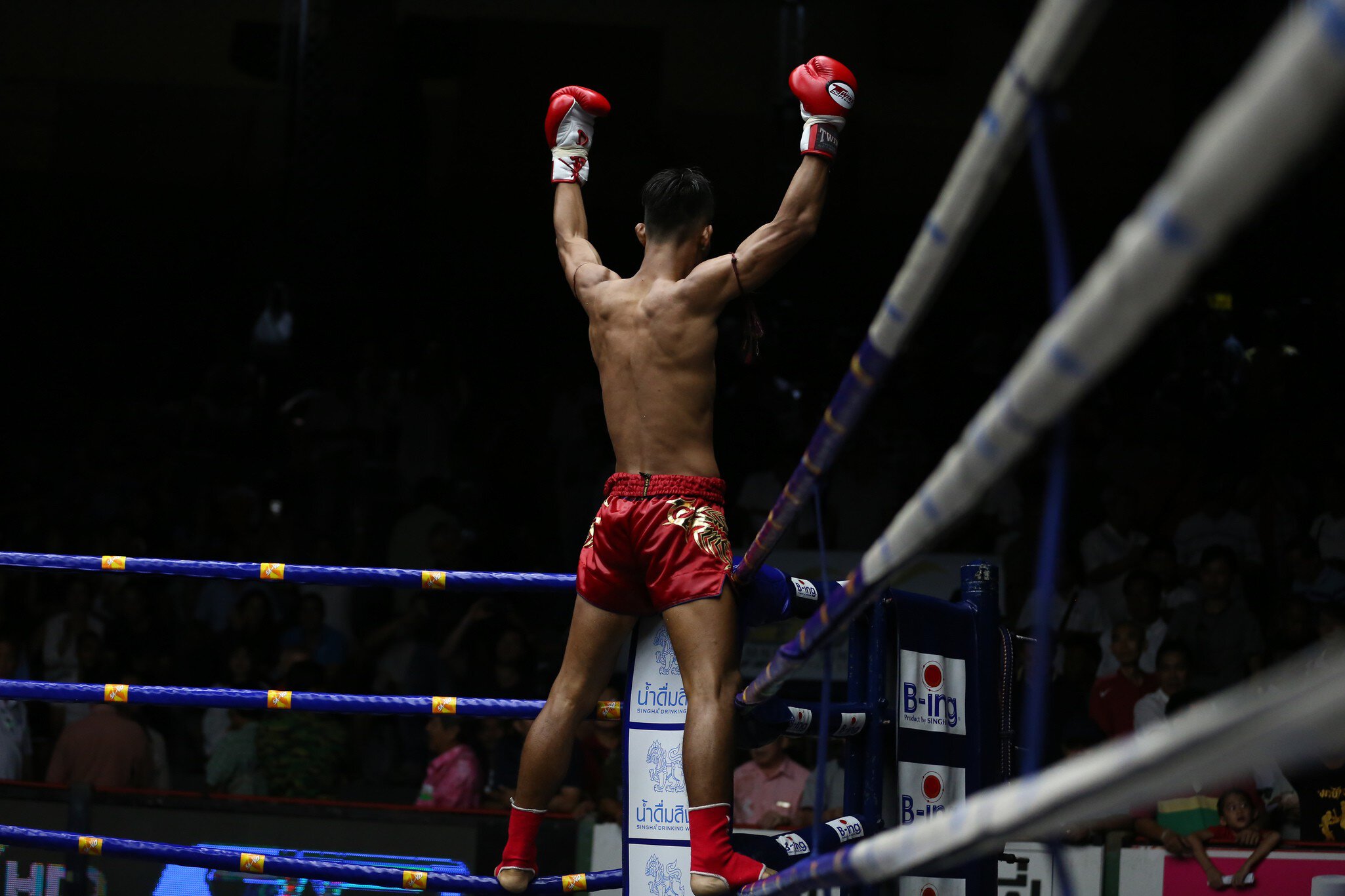 A fighter stands on the ropes of a boxing ring with his arms up after a fight