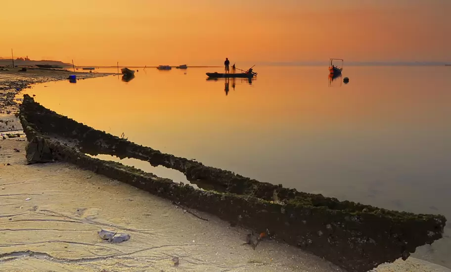 Fisherman stands on a small boat on calm water at orange sunset off Johor Bahru, a rotting wooden hull on the sand