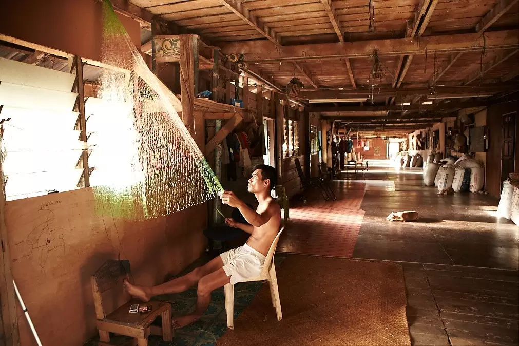 A man sits on a chair in a long corridor repairing a fishing net, in Batang Ai National Park.