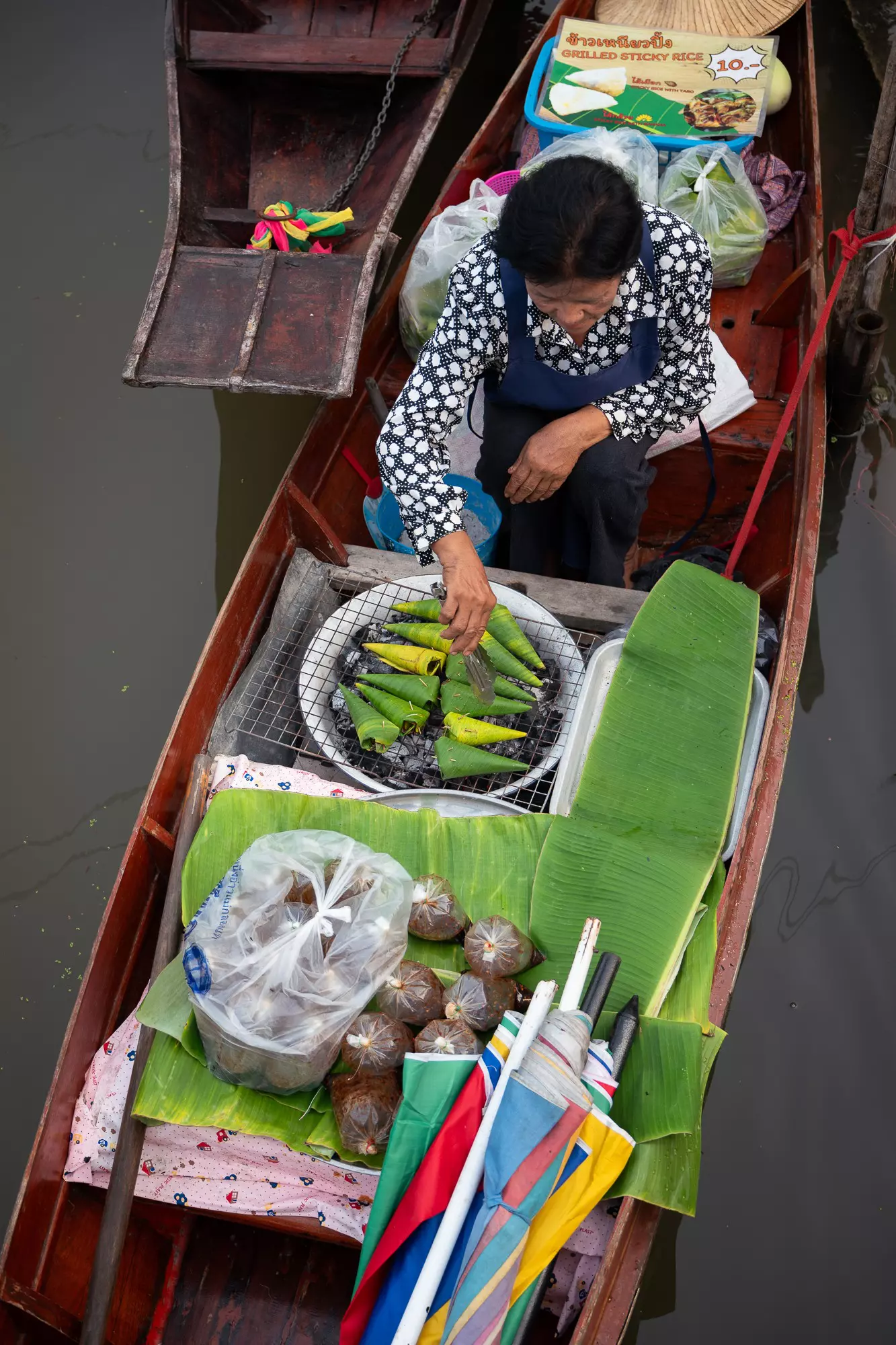 An overhead view of a woman grilling food wrapped in banana leaves on a boat in a floating market.
