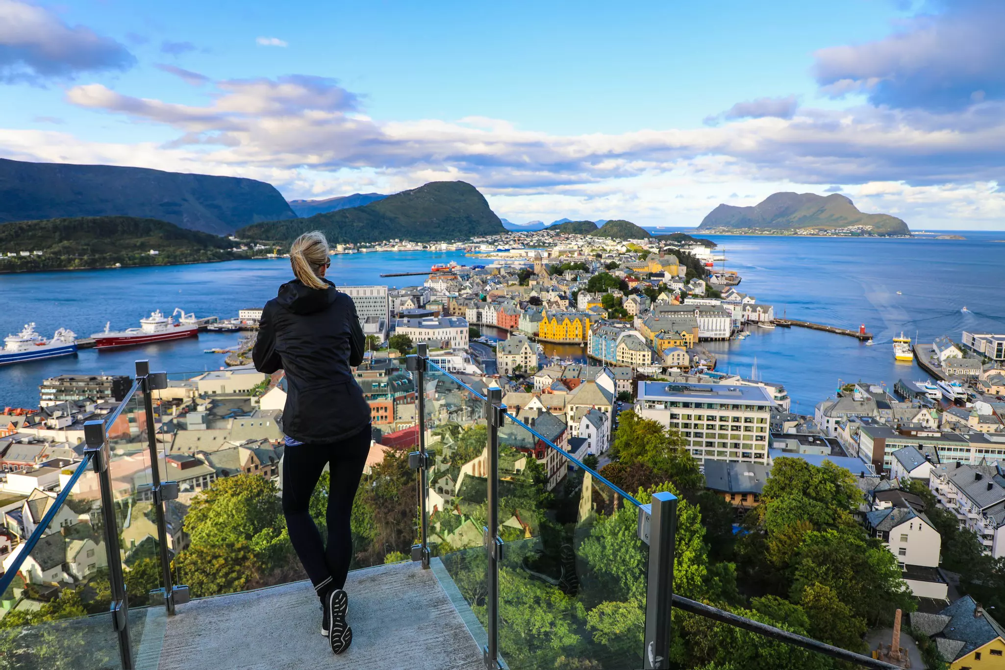 A woman is seen from behind at a viewpoint overlooking a city surrounded by water and distant hills.