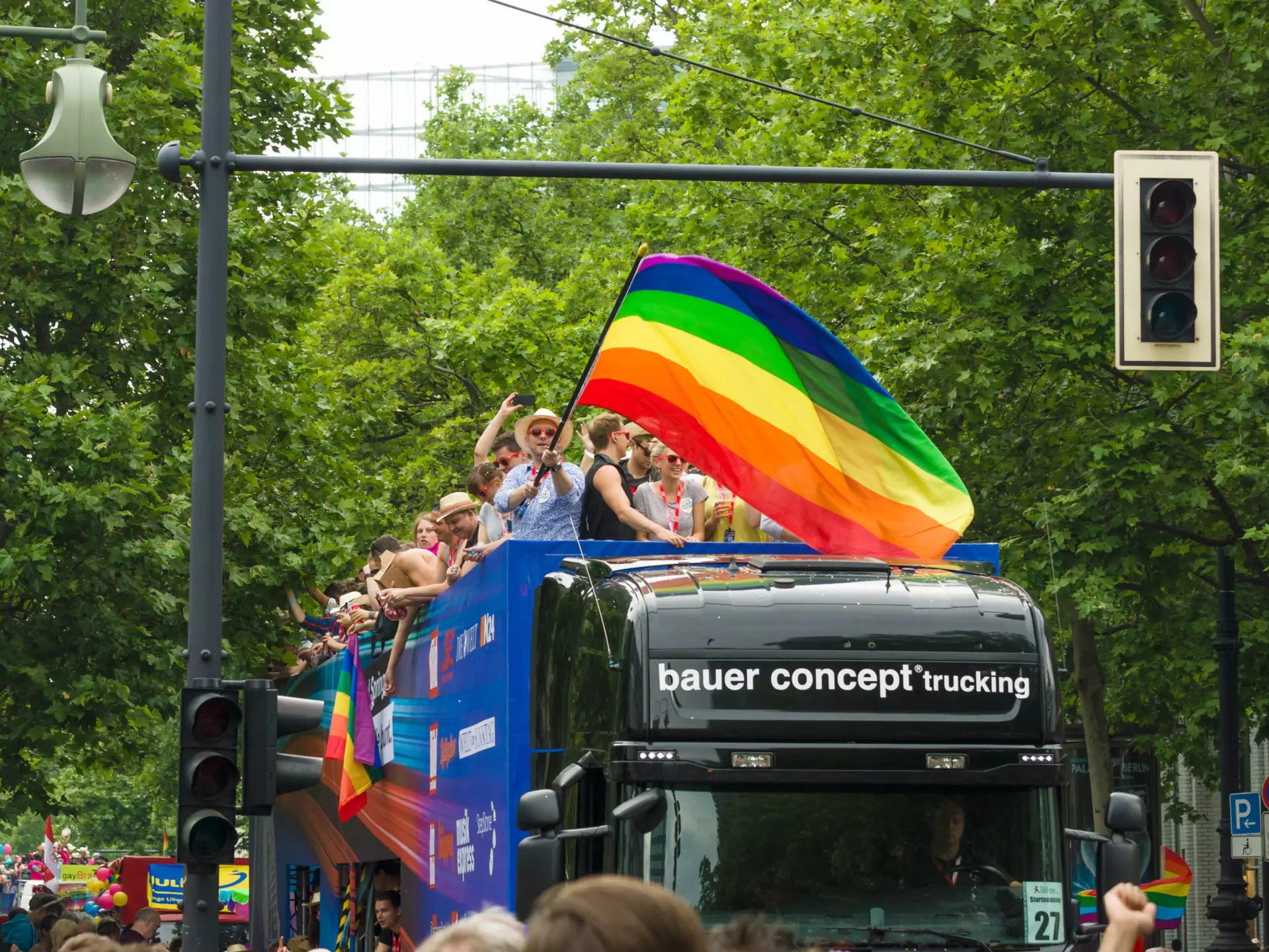 People waving a rainbow flag stand on the top deck of a black bus making its way down a street