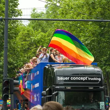 People waving a rainbow flag stand on the top deck of a black bus making its way down a street