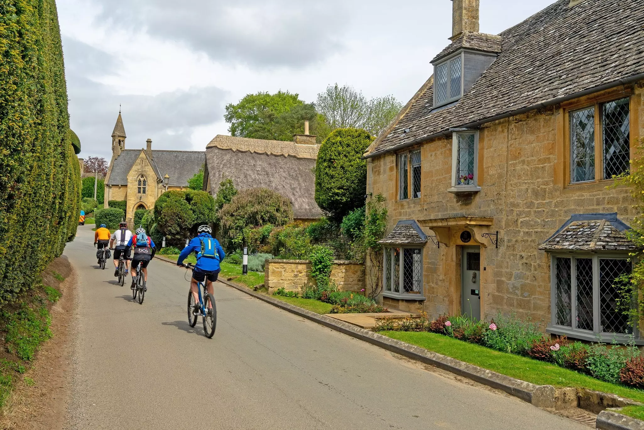 Four cyclists peddle through a village with thatched cottages and a quaint church.