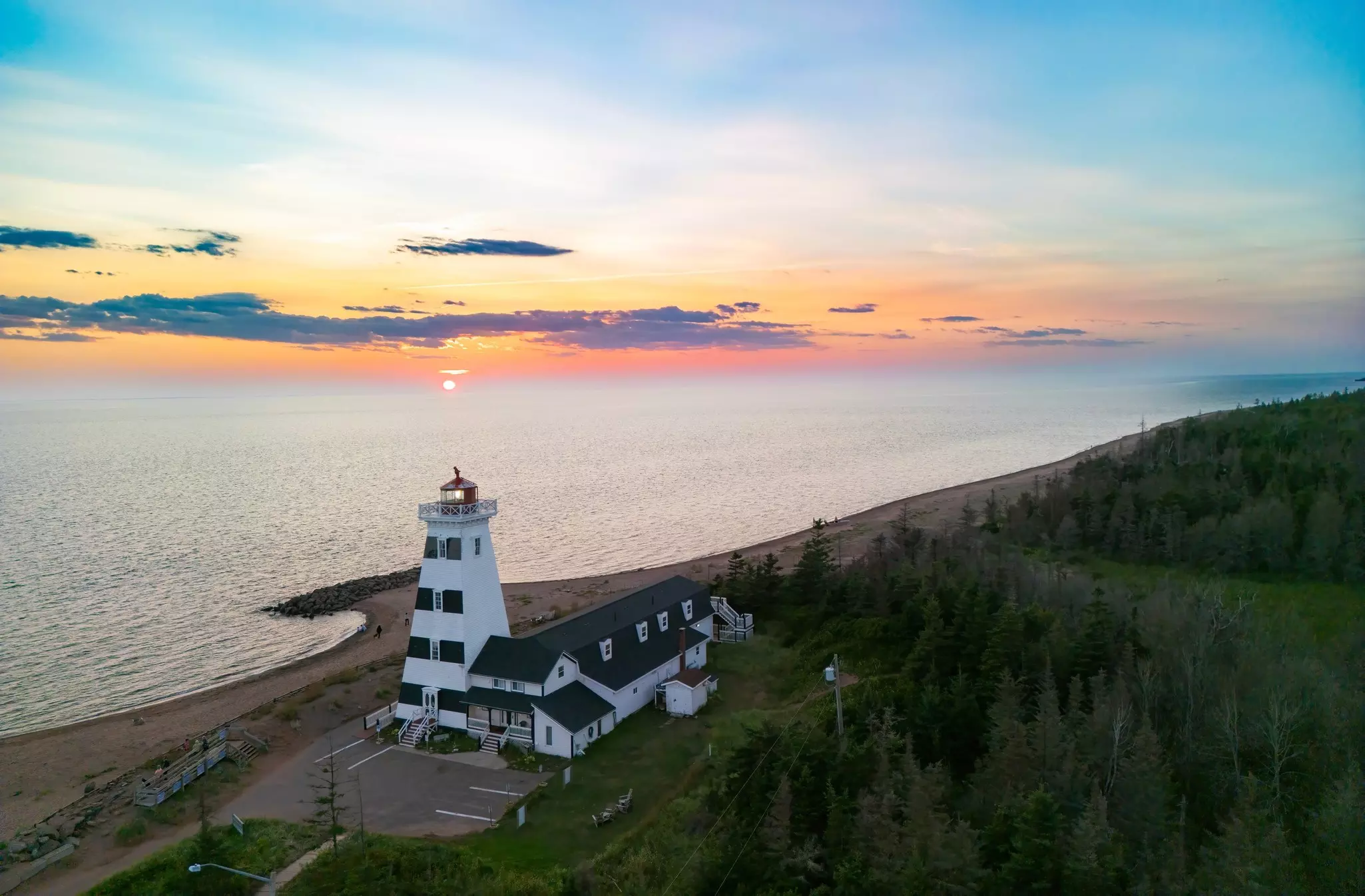 A white and black lighthouse on the shore as the sun sets over the calm ocean waters.