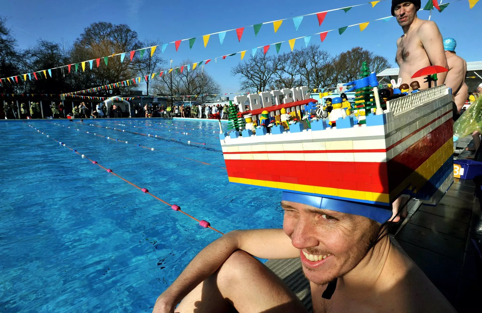 A competitor wears a fancy hat made of plastic bricks at an outdoor pool.