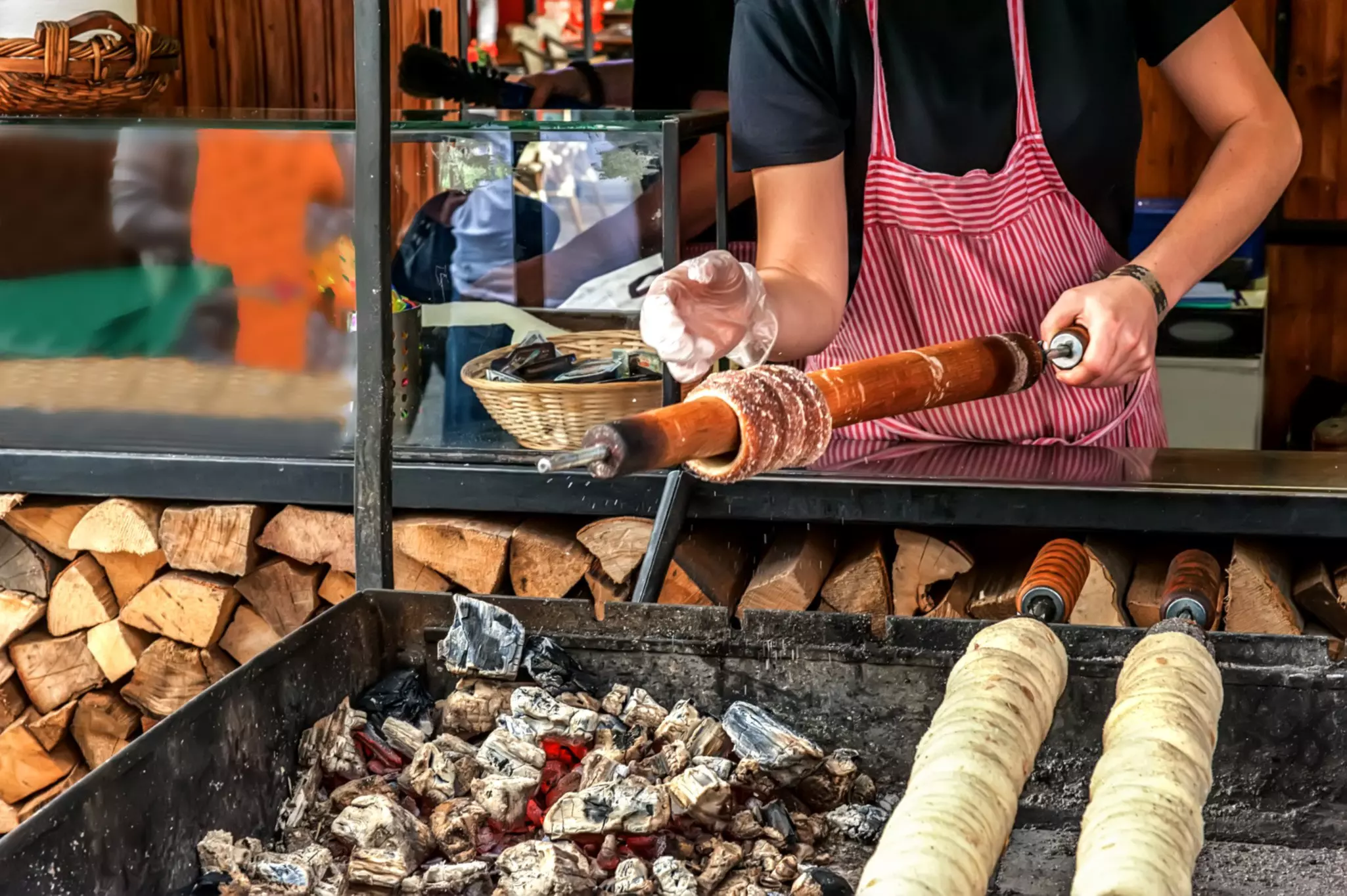 A person wearing a red striped apron holds a bar with dough wrapped around it over hot coals.