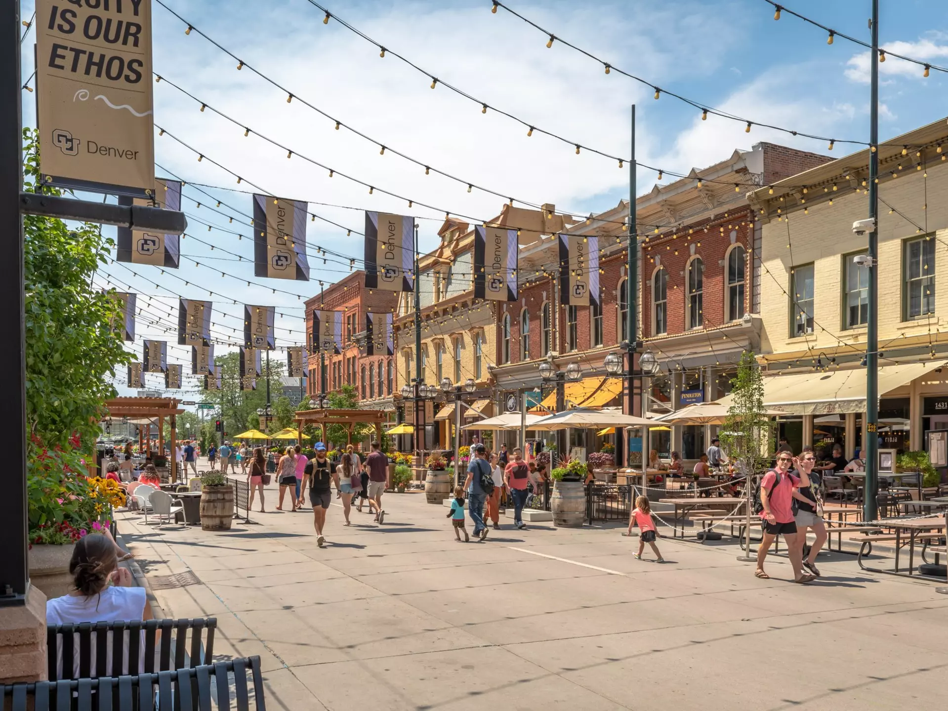People walking on pedestrian streets lined with restaurants and shops on a sunny day.