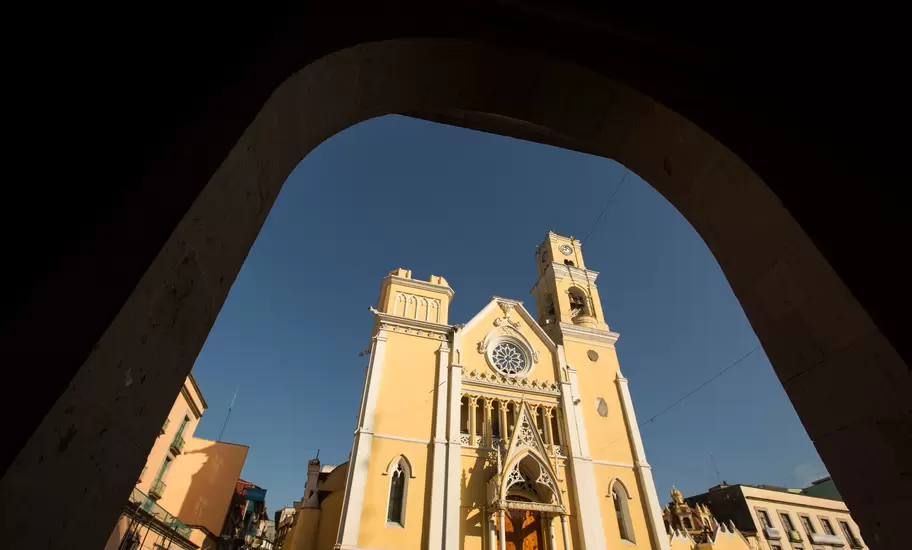 Yellow neo-Gothic facade and clock tower of Xalapa Cathedral, Veracruz, framed by a dark stone archway against blue sky