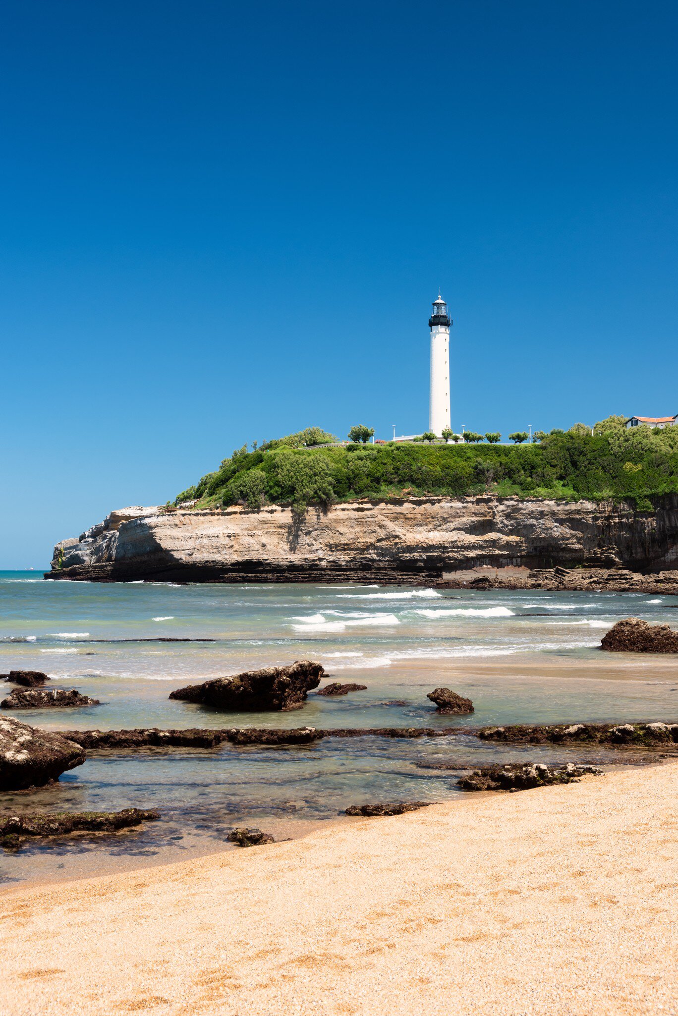 A tall white lighthouse on a green clifftop above layered rock strata, with rocks scattered across the sand and shallows below.