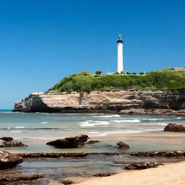 A tall white lighthouse on a green clifftop above layered rock strata, with rocks scattered across the sand and shallows below.