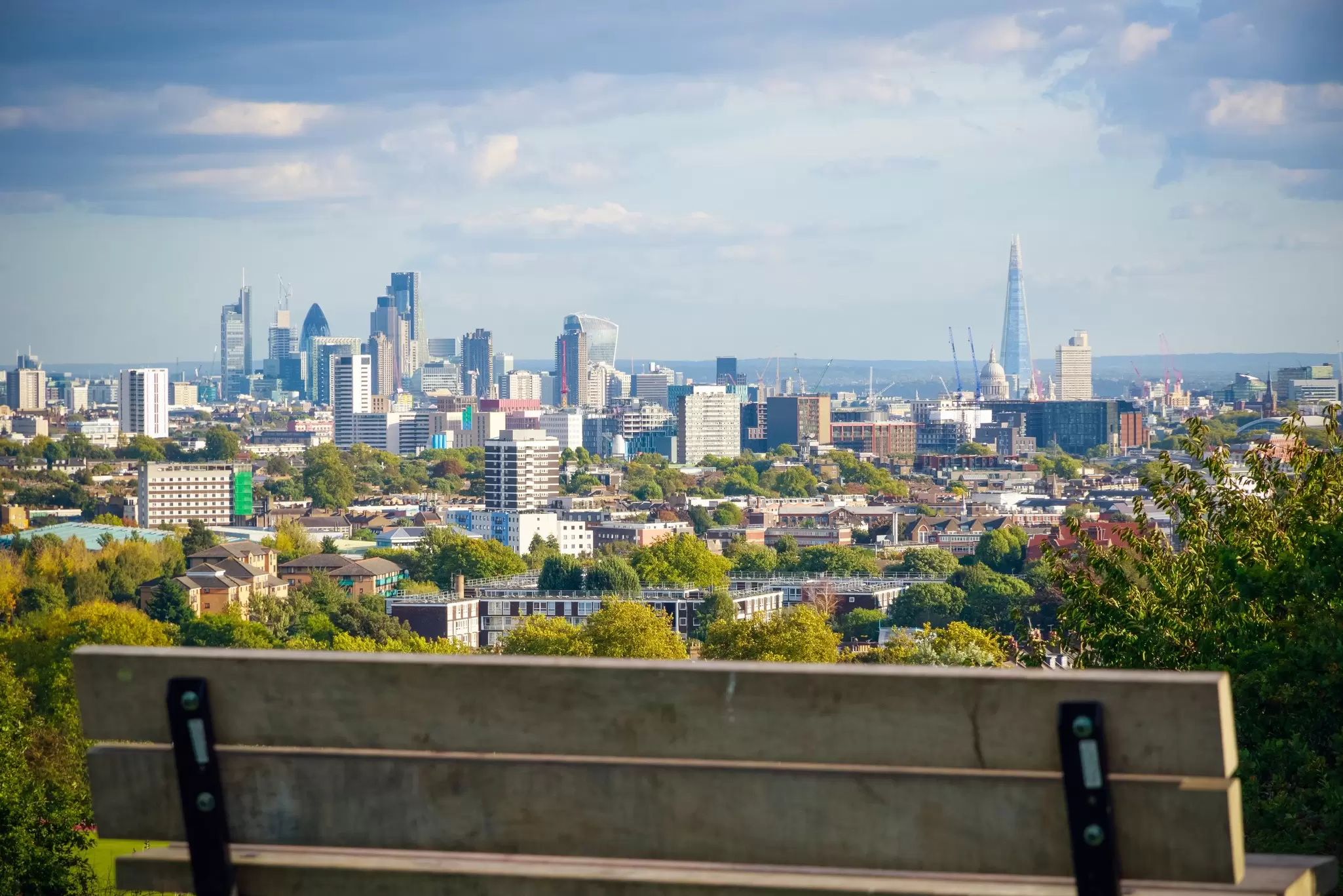 An empty bench on Primrose Hill with a distant view of skyscrapers and tall buildings in the center of London.