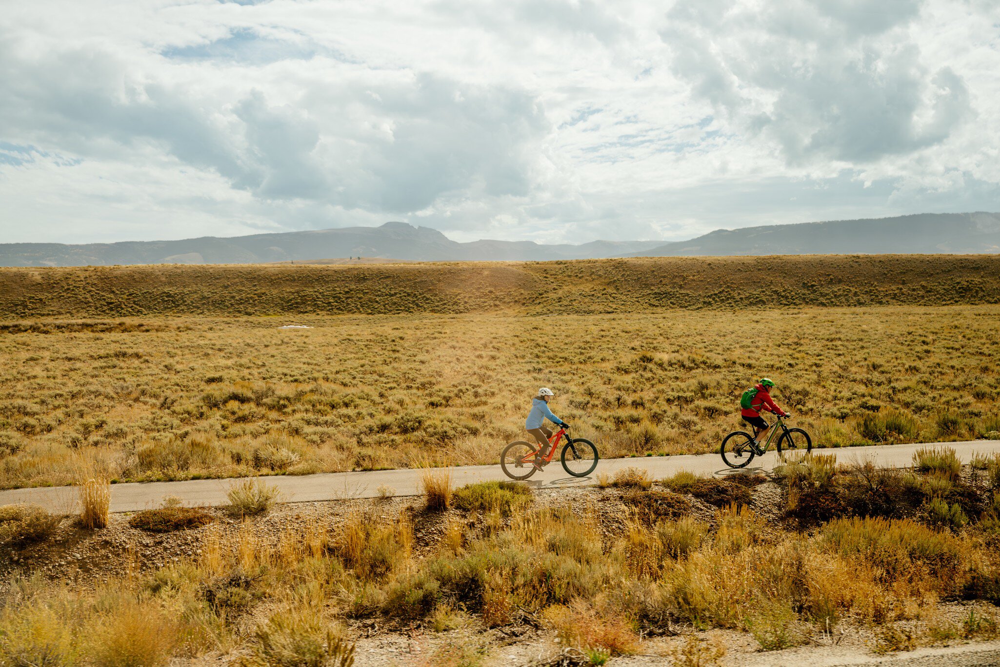 Two cyclists ride a paved pathway through sagebrush flats in Grand Teton National Park under heavy clouds