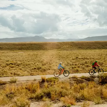 Two cyclists ride a paved pathway through sagebrush flats in Grand Teton National Park under heavy clouds