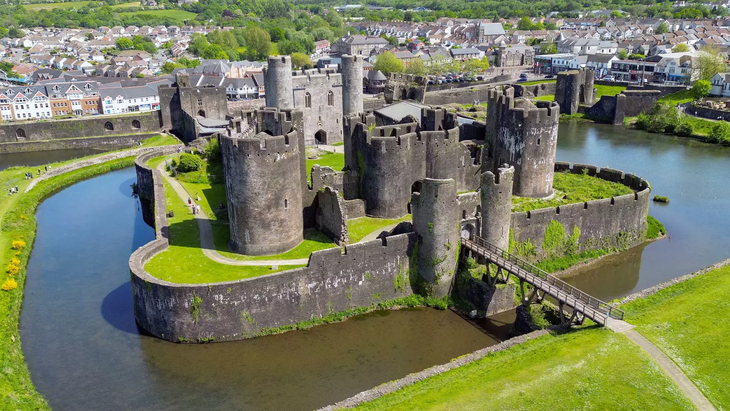 Castle towers surrounded by a moat and bright green grass, with homes and trees in the distance on a sunny day.