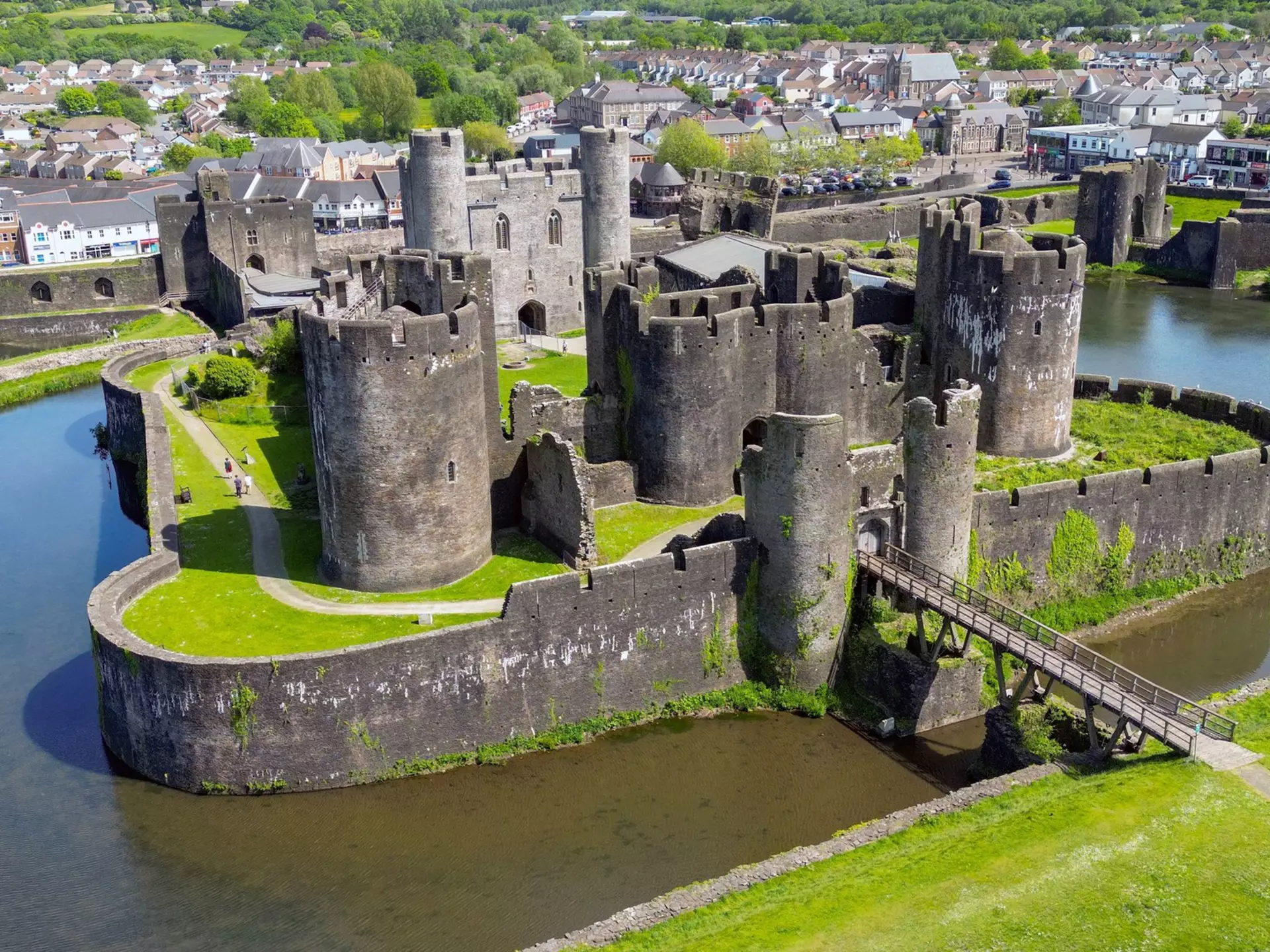 Castle towers surrounded by a moat and bright green grass, with homes and trees in the distance on a sunny day.