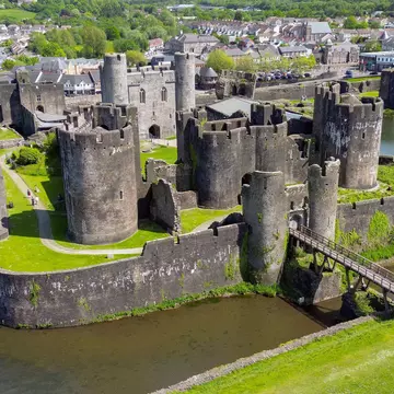 Castle towers surrounded by a moat and bright green grass, with homes and trees in the distance on a sunny day.