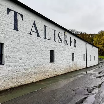 A long whitewashed distillery warehouse with TALISKER painted in tall dark letters along its wall, beside a rain-soaked road.