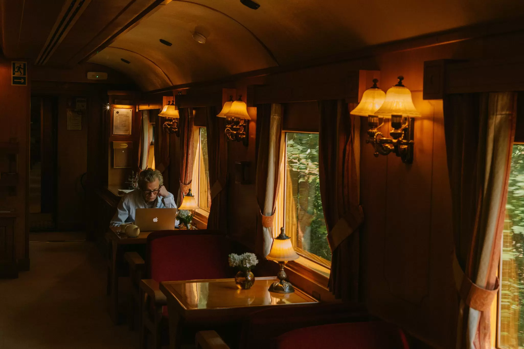 A guest in the lounge of a swanky train making use of wifi onboard on her laptop. The walls are made of glossy wood and the furniture is luxurious and plush with low-lit wall sconces.
