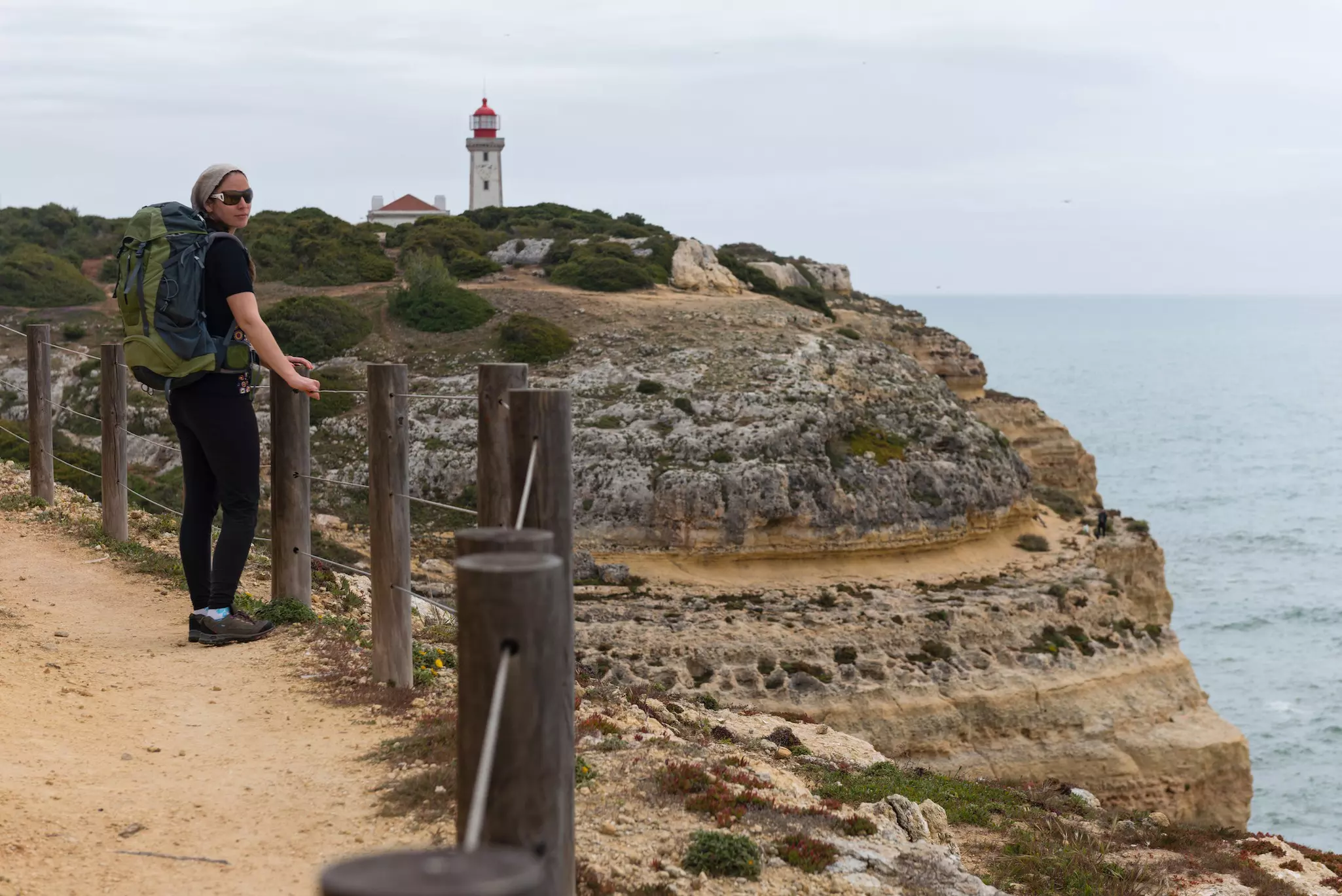 A hiker standing on a path on the edge of a cliff overlooking water, with a white and red lighthouse in the distance.