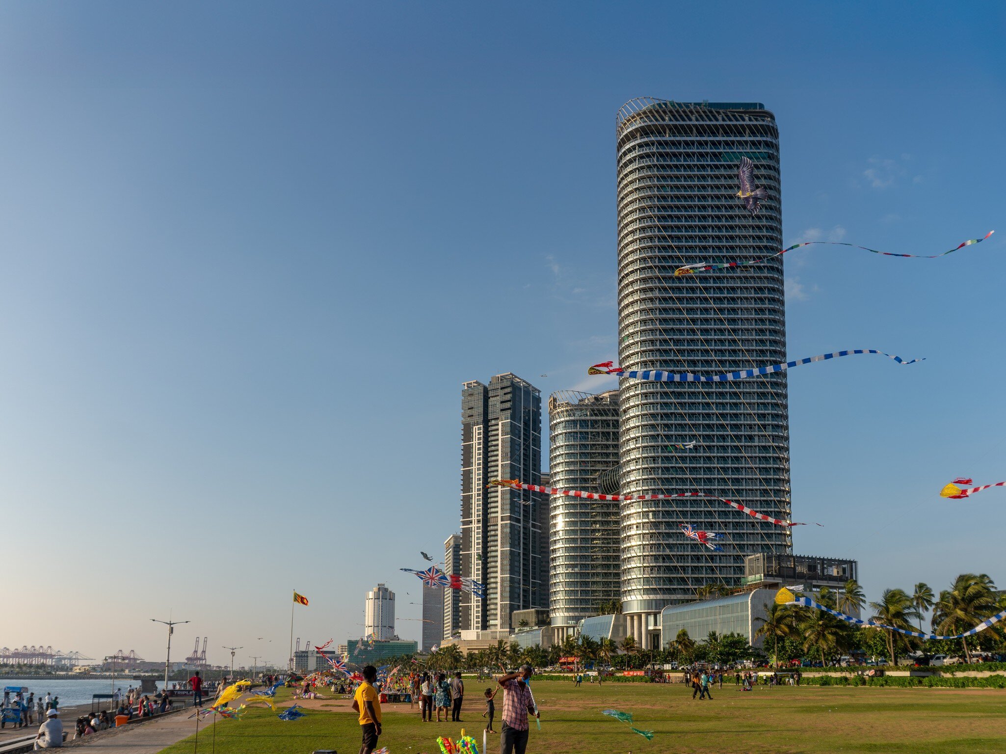 People on a wide seafront lawn below glass towers, with kites flying above.