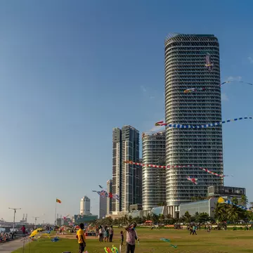People on a wide seafront lawn below glass towers, with kites flying above.