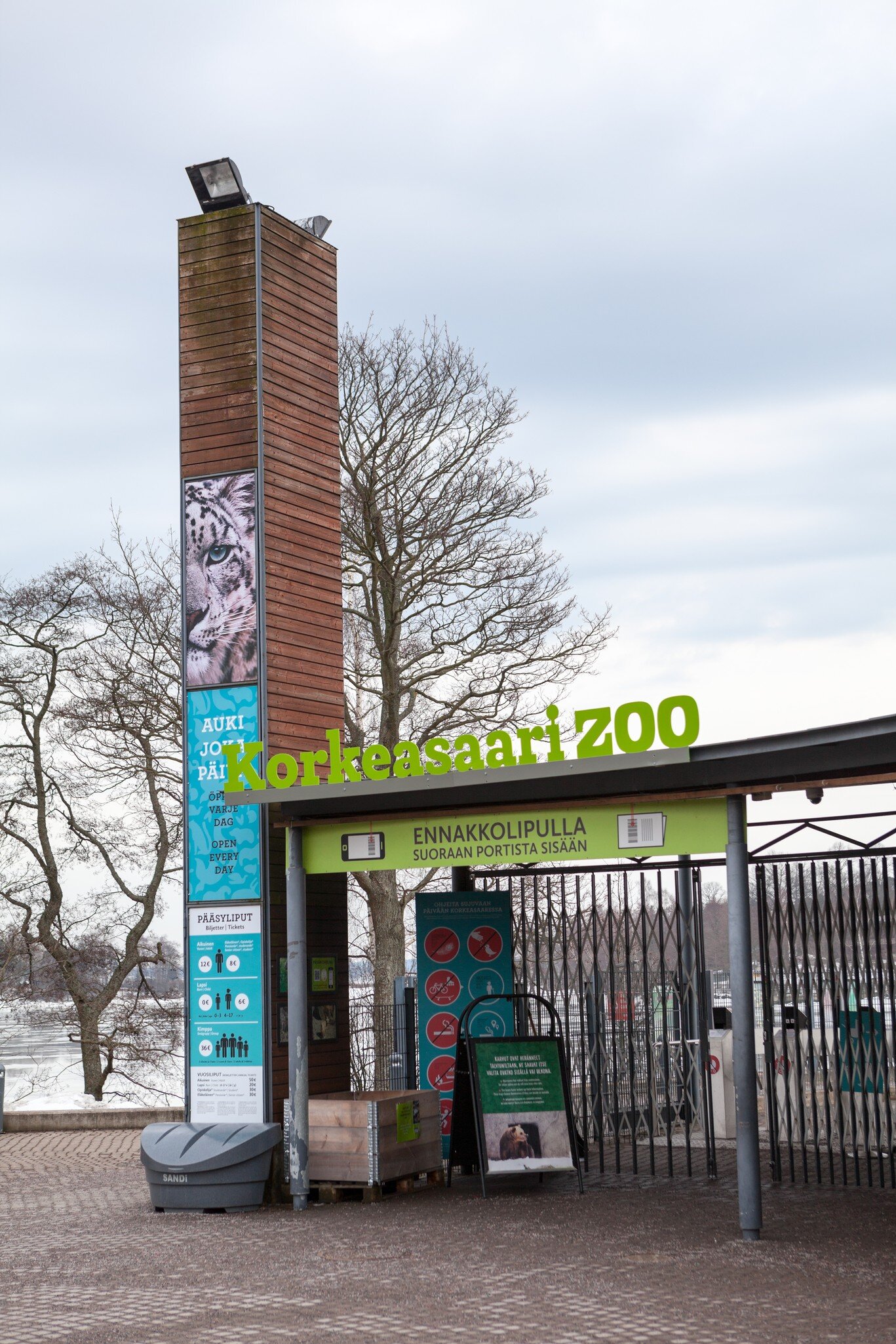 Green 'Korkeasaari ZOO' lettering over closed entry gates, beside a timber pylon with a snow leopard poster and ticket prices