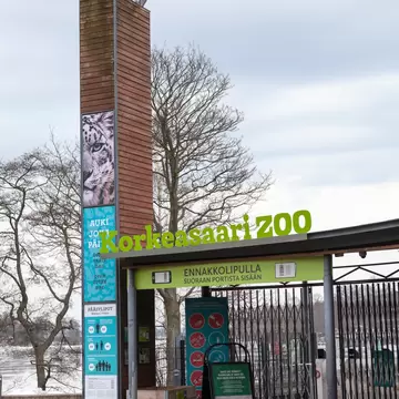 Green 'Korkeasaari ZOO' lettering over closed entry gates, beside a timber pylon with a snow leopard poster and ticket prices