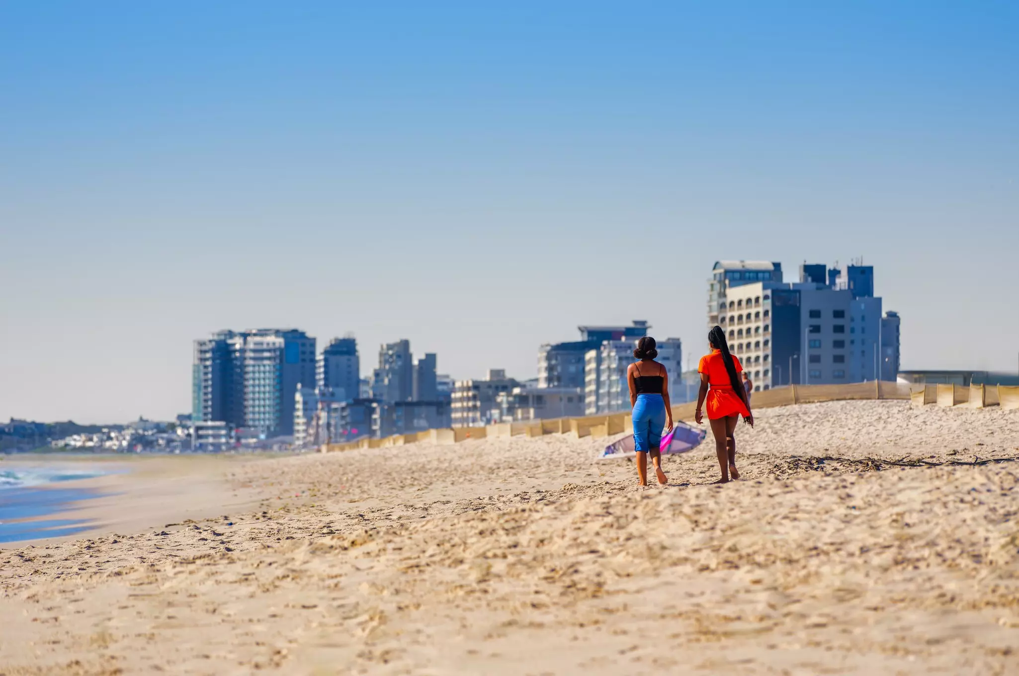 Two young women walk along a sandy beach backed by high-rise apartment buildings.